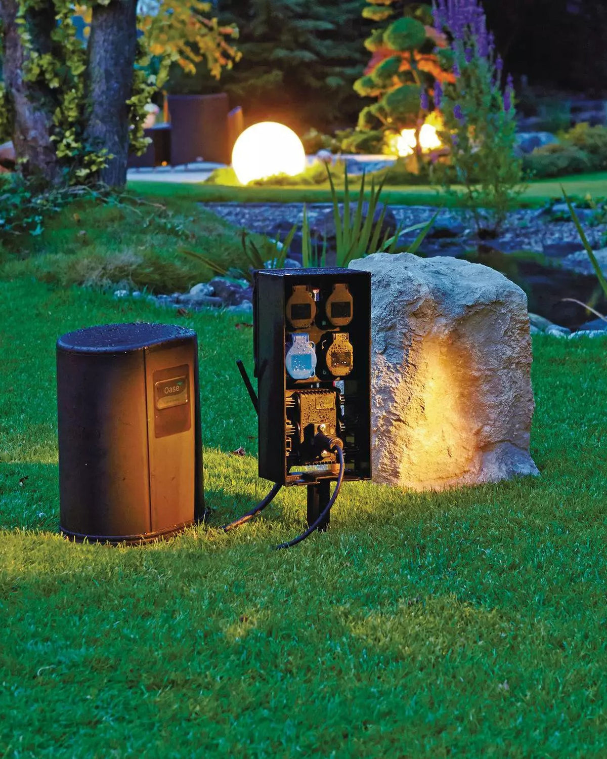 OASE InScenio FM-Master Cloud am Abend im Garten, daneben Haube und ein beleuchteter Stein