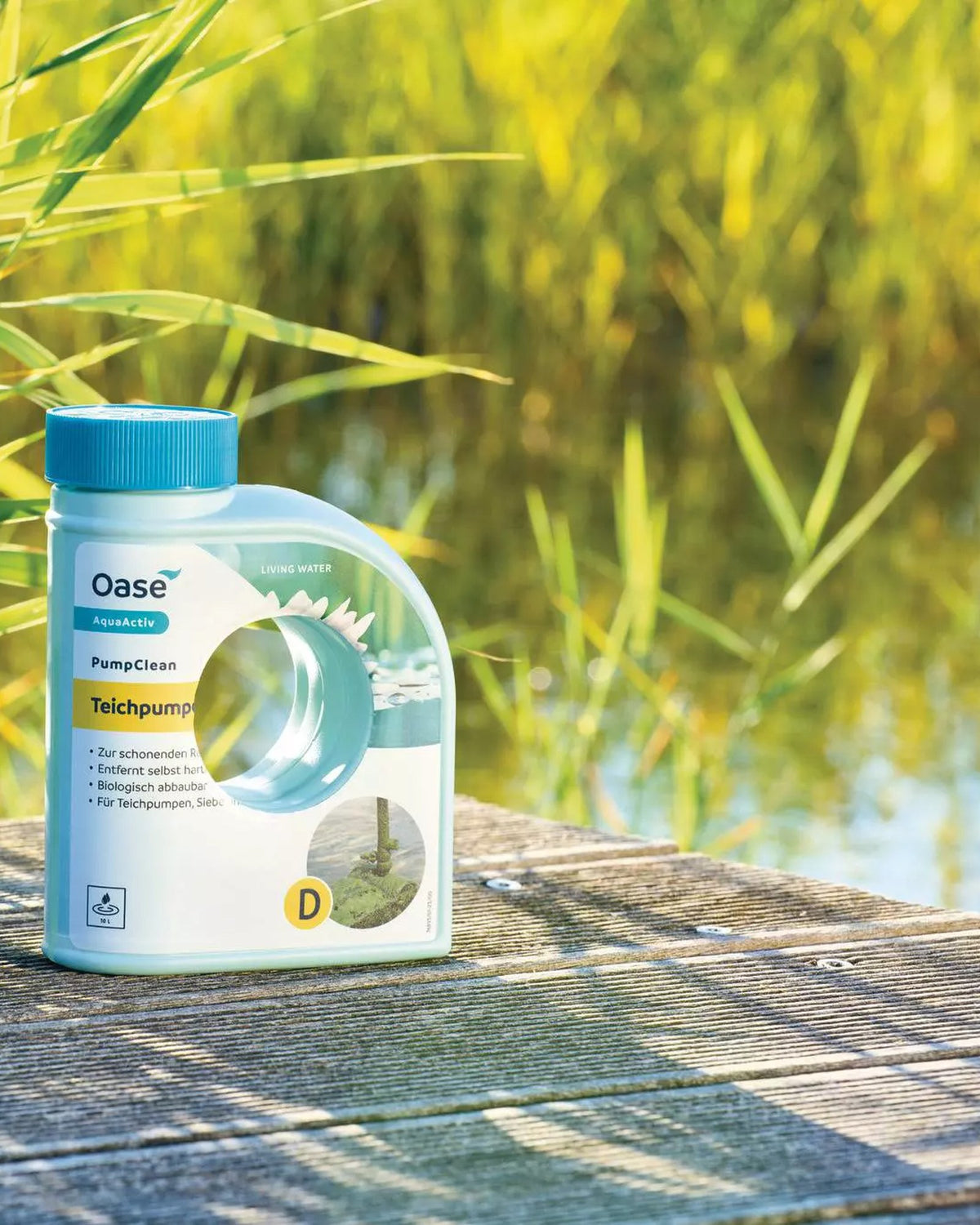 OASE AquaActiv PumpClean 500 ml auf einem Holzsteg am Teich, im Hintergrund Schilf